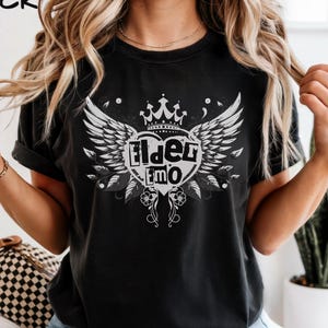 Pode incluir: Camiseta preta com um design gráfico branco com uma coroa, asas e o texto "Elder Emo". A palavra "BLACK" está impressa no canto superior esquerdo. A camisa é um item de roupa casual.