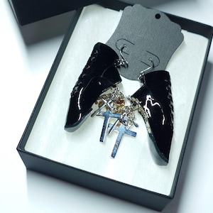 以下が含まれることがあります： シルバーのクロスチャームと小さな金色のビーズが付いた、光沢のある黒いイヤリング。 イヤリングは、白い内装の黒いギフトボックスと黒いイヤリングディスプレイカードに表示されています。