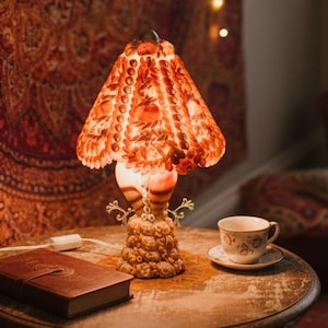May include: A vintage-style table lamp with an ornate base and a patterned lampshade. The lamp sits on a round wooden table with a book and teacup. A tapestry and string lights are in the background, creating a warm, inviting atmosphere.