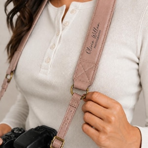 May include: A dusty rose camera strap featuring the text Olivia Willam PHOTOGRAPHY in black script. The strap is constructed with stitched leather-like material, antique brass-toned metal rings, and adjustable buckles attached to a black camera.