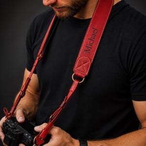May include: A red leather camera strap featuring visible stitching and brass-toned metal hardware. The strap is personalized with the name Michael embossed in a dark font. It connects to a black camera body via thin leather loops and metal rings.