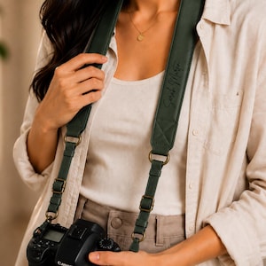 May include: A dark forest green camera strap featuring a wide padded section with the name Sarah Martin printed in black script. The strap includes antique brass-coloured metal hardware, adjustable buckles, and circular rings connecting to a black camera.