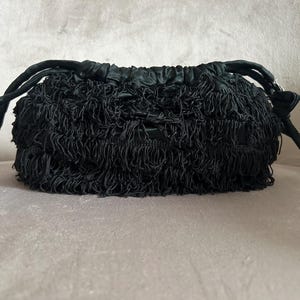Può includere: Una borsa nera con coulisse e esterno strutturato e frangiato. La borsa presenta una parte superiore arricciata e manici in corda neri. Il materiale frangiato conferisce alla borsa un aspetto tattile unico. La borsa è su una superficie rosa chiaro.