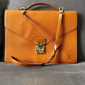 May include: A tan leather briefcase featuring a textured horizontal line pattern, a top handle, and a detachable shoulder strap. The bag includes a gold-tone metal clasp closure with an embossed logo and a small LV logo stamped in the bottom corner.