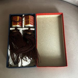 May include: A rectangular box with a dark blue interior and a red lid containing two small ceramic ornaments. Each ornament features a red and white geometric pattern with gold accents and is attached to a long, dark brown decorative tassel.