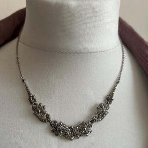 Puede incluir: Un collar plateado con un diseño ornamentado y simétrico. El collar presenta un motivo central en forma de flor y patrones más pequeños y repetidos a lo largo de la cadena. El collar se exhibe en un busto de maniquí blanco.