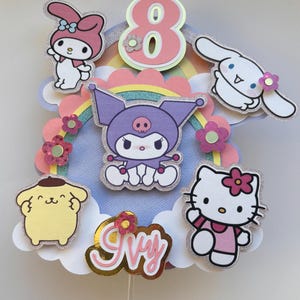May include: A vibrant cake topper with cartoon characters, a rainbow, and the number 8. The topper includes characters like Hello Kitty and My Melody, with the name "Ivy" in gold script. The design is set against a blue background.