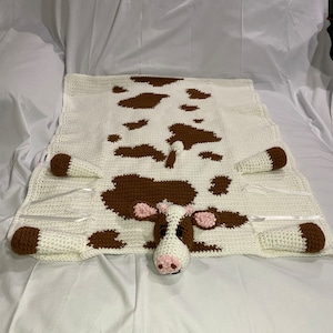 May include: A crocheted cow blanket with brown spots on a white background. The cow has a pink nose and ears.