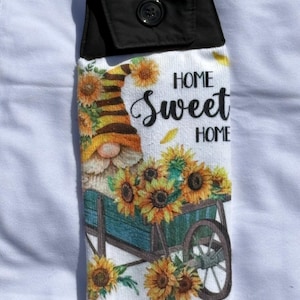 May include: A hanging kitchen towel with a black top and button closure. The towel features a gnome pushing a wheelbarrow filled with sunflowers. The words "HOME Sweet HOME" are printed on the towel.