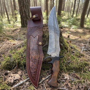 Può includere: Un grande e ornato coltello da caccia con una lama a motivi e un manico in legno, accanto a un fodero in pelle marrone. Il fodero presenta un motivo a pelle di coccodrillo e la parola "VIKING" è visibile. Il coltello è ambientato in un bosco.