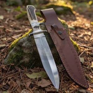 May include: A hunting knife with a polished steel blade and an antler handle. The knife is next to a brown leather sheath, resting on a mossy rock. The blade features an etched design. The background is a natural outdoor setting.