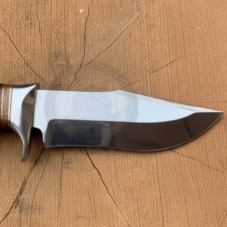 Peut inclure: Gros plan sur un couteau de chasse avec une lame en acier poli et un manche marron et argent&eacute;. La lame a un bord incurv&eacute; et une pointe. Le manche est en bois et en m&eacute;tal. Le couteau est sur une surface en bois.
