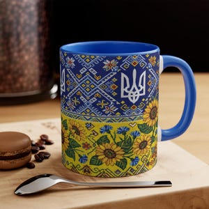 May include: A white ceramic mug with a black handle and the text "But First, Coffee" in a bold, black font. The mug is set against a blurred background, suggesting a cozy setting.
