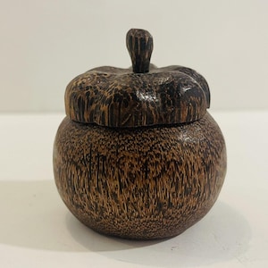 May include: A small, round wooden container with a lid. The container and lid are a dark brown colour with a textured wood grain pattern. The lid has a small, carved stem-like handle. The container appears to be handmade.