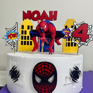 May include: A white cake decorated with a Spider-Man theme. The cake features a Spider-Man figure, the name "NOAH" in red letters, and a number "4" with comic book-style accents. The cake also has a Spider-Man mask and spiderweb decorations.