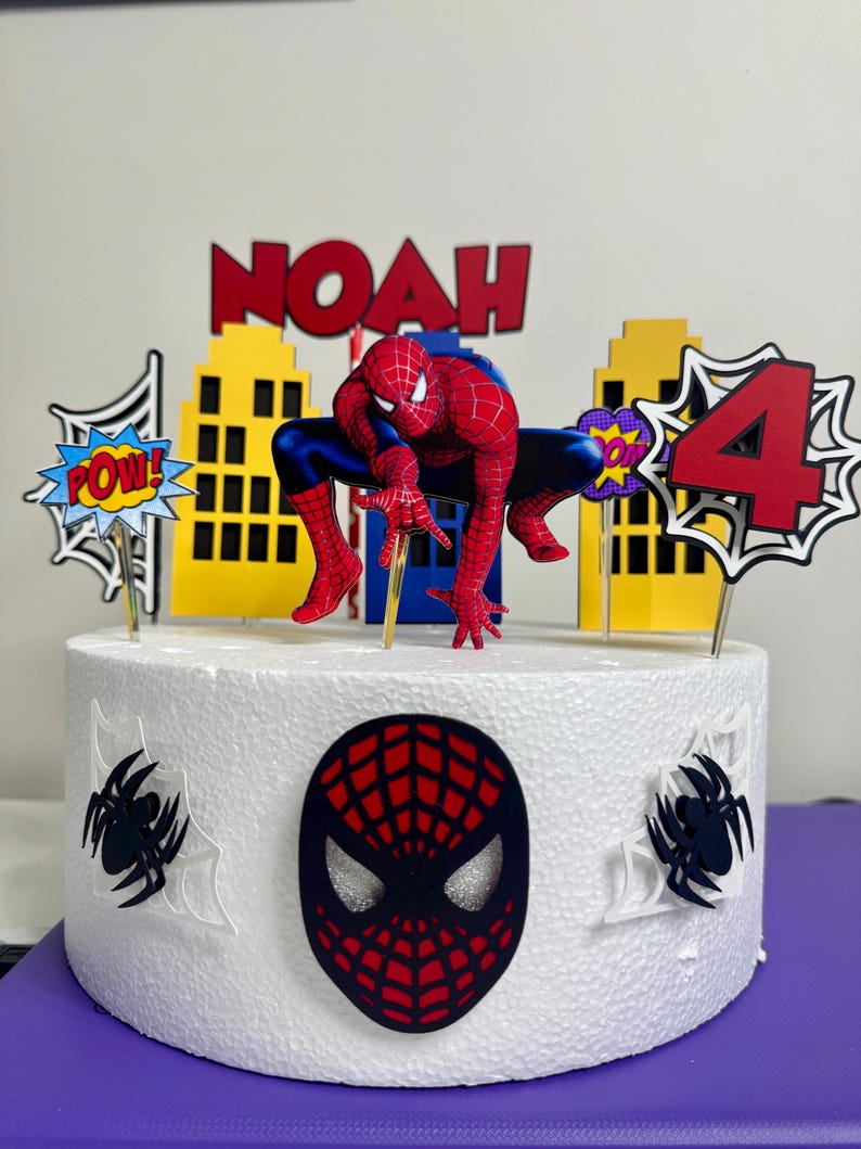 May include: A white cake decorated with a Spider-Man theme. The cake features a Spider-Man figure, the name "NOAH" in red letters, and a number "4" with comic book-style accents. The cake also has a Spider-Man mask and spiderweb decorations.