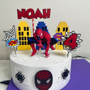 May include: A round white cake decorated with a Spider-Man theme. The cake features a Spider-Man figurine, cityscape cutouts, and the name "NOAH" in red letters. Additional decorations include a "4" and comic book style speech bubbles.