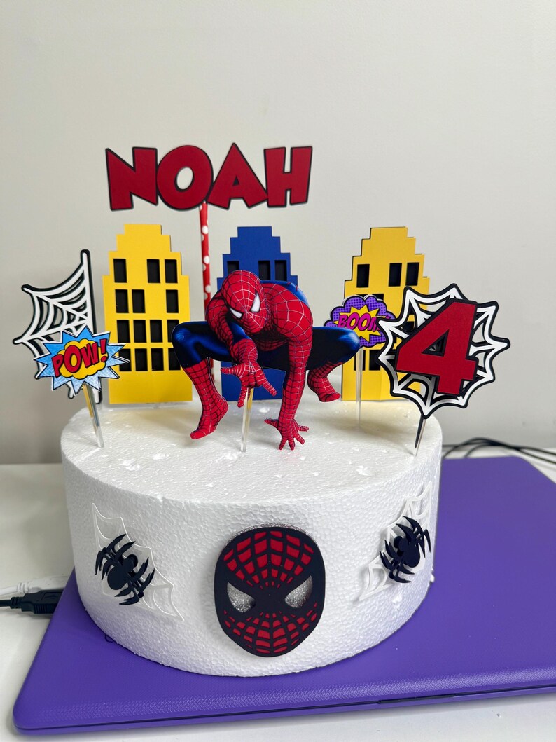 May include: A round white cake decorated with a Spider-Man theme. The cake features a Spider-Man figurine, cityscape cutouts, and the name "NOAH" in red letters. Additional decorations include a "4" and comic book style speech bubbles.