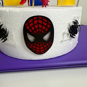 May include: A white cake decorated with a black and red Spider-Man mask, spiderwebs, and a cityscape. The cake features red and blue Spider-Man figures and yellow and blue building cutouts. The cake is on a purple surface.