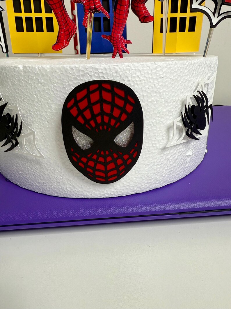 May include: A white cake decorated with a black and red Spider-Man mask, spiderwebs, and a cityscape. The cake features red and blue Spider-Man figures and yellow and blue building cutouts. The cake is on a purple surface.