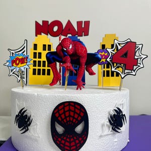 May include: A white cake decorated with a Spider-Man theme. The cake features a Spider-Man figure, the name "NOAH" in red letters, and a number "4" with comic book-style accents. The cake is adorned with spider web and spider decorations.