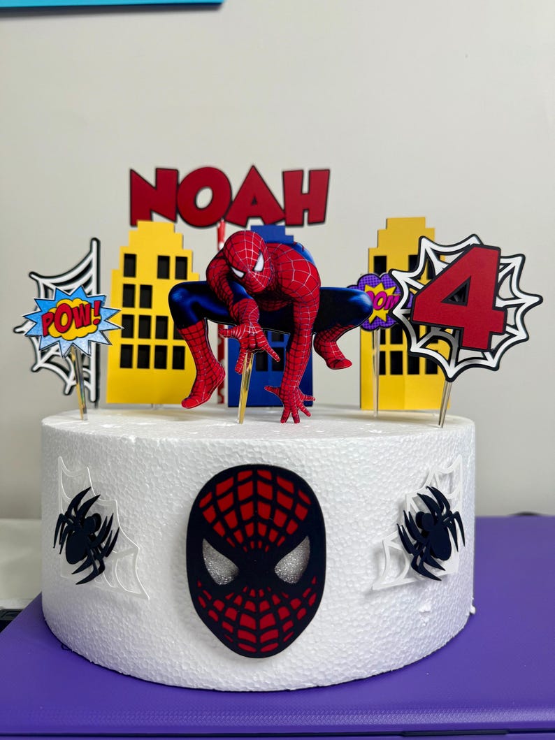 May include: A white cake decorated with a Spider-Man theme. The cake features a Spider-Man figure, the name "NOAH" in red letters, and a number "4" with comic book-style accents. The cake is adorned with spider web and spider decorations.