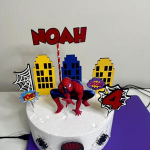 May include: A round white cake decorated with a Spider-Man theme. It features a Spider-Man figurine, cityscape cutouts, and the name "NOAH" in red. The cake also has a "4" and comic book sound effects.