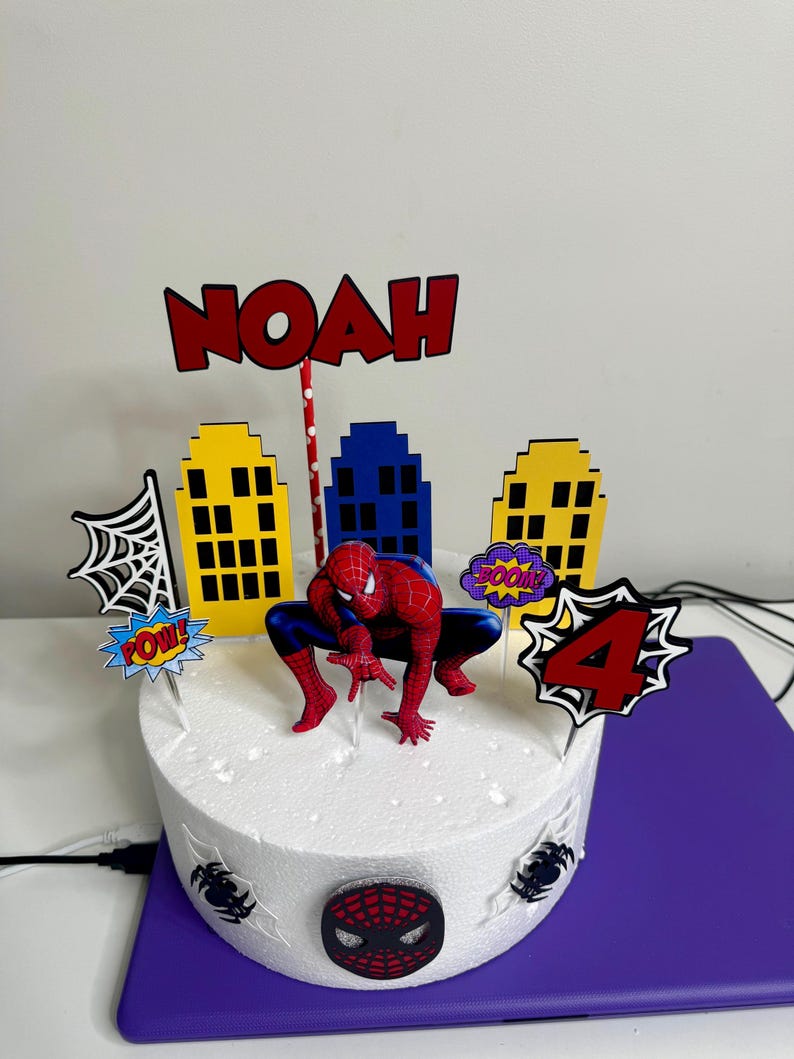 May include: A round white cake decorated with a Spider-Man theme. It features a Spider-Man figurine, cityscape cutouts, and the name "NOAH" in red. The cake also has a "4" and comic book sound effects.