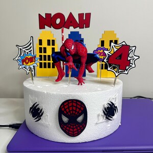 May include: A round white cake decorated with a Spider-Man theme. The cake features a Spider-Man figurine, cityscape cutouts, and the name "NOAH" in red letters. The number "4" is also displayed, along with comic book style speech bubbles.