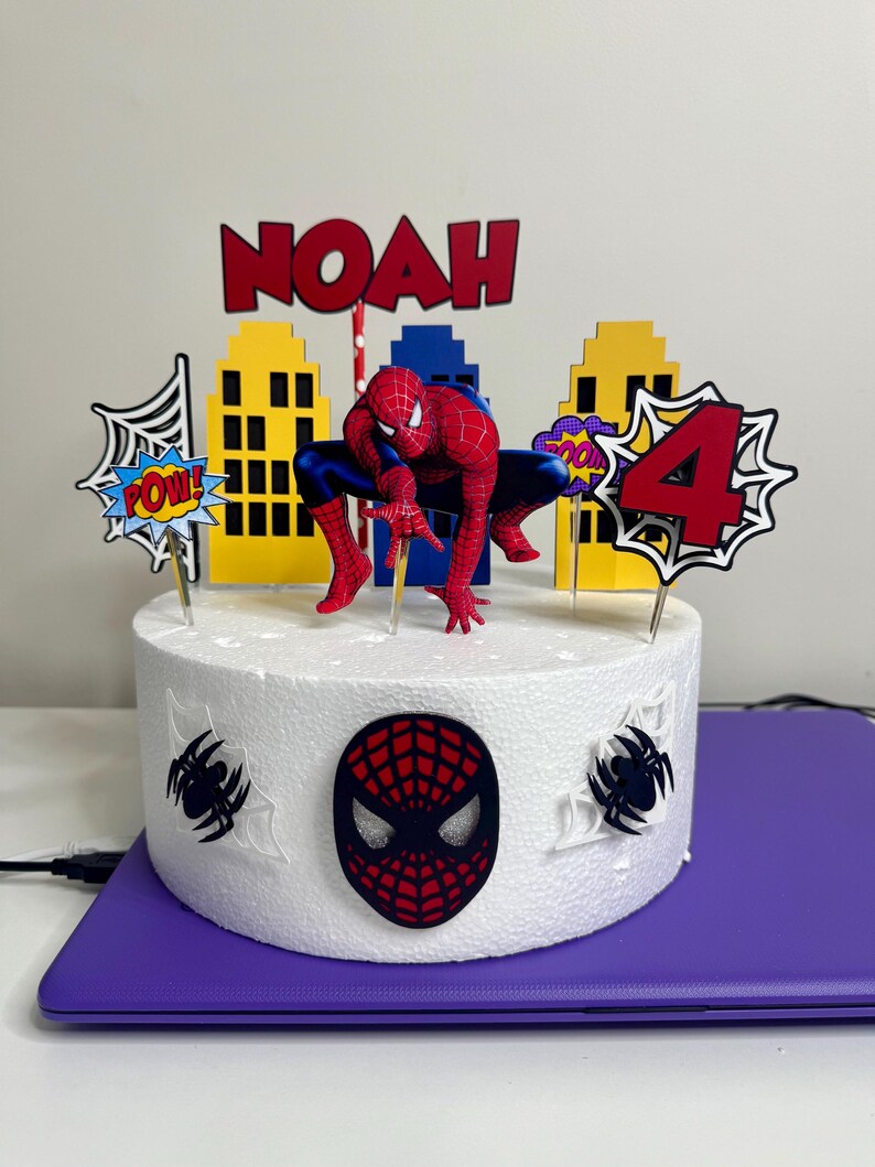 May include: A round white cake decorated with a Spider-Man theme. The cake features a Spider-Man figurine, cityscape cutouts, and the name "NOAH" in red letters. The number "4" is also displayed, along with comic book style speech bubbles.