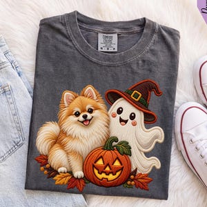 May include: A charcoal gray t-shirt featuring a graphic of a fluffy tan Pomeranian dog sitting next to a white ghost wearing a brown witch hat and a carved orange jack-o'-lantern pumpkin surrounded by colorful autumn leaves on the front.