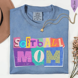 May include: A light blue Comfort Colors t-shirt with the words "Softball Mom" in colorful, cut-out style letters. A gold star pendant necklace rests on the shirt. The shirt is folded and placed on a white surface.