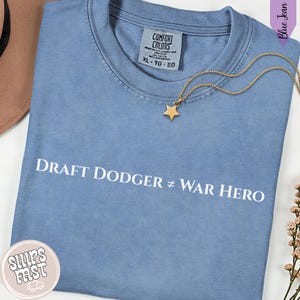 May include: A light blue t-shirt with the text "DRAFT DODGER ≠ WAR HERO" in white. A gold star pendant hangs on a gold chain. The shirt has a Comfort Colors tag, size XL.