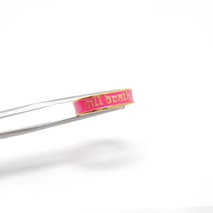 Op de afbeelding: Een goudkleurige ring met een felroze band. De ring heeft de woorden "till death" in een gotisch lettertype. De ring wordt vastgehouden door een zilveren pincet tegen een witte achtergrond.