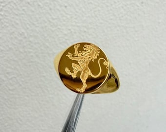 May include: A gold-toned signet ring featuring a circular face with an engraved lion design. The lion is depicted in a rampant position, standing on one hind leg with its tail curved upward. The ring has a polished, reflective metallic surface.