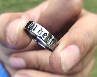 Op de afbeelding: Een zilveren ring met de woorden "till death" gegraveerd op de band. De ring wordt tussen de vingers gehouden, waarbij de tekst duidelijk zichtbaar is. De ring heeft een gepolijste afwerking en een klassiek ontwerp.