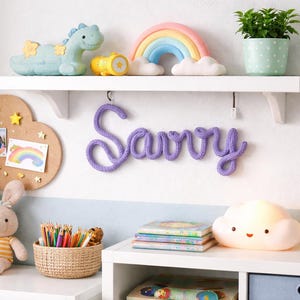 May include: A child's bedroom scene featuring a white shelf with a plush dinosaur, rainbow, and plant. A purple 'Savvy' name sign hangs below. A cloud lamp, bunny, and corkboard with photos are on a white shelf.