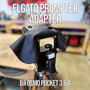 May include: Black Elgato Prompter Adapter for DJI Osmo Pocket 3 & 4. The adapter features a black frame with a black fabric hood. The DJI Osmo Pocket camera is mounted on the adapter, displaying its screen. The text "ELGATO PROMPTER ADAPTER" and "DJI OSMO POCKET 3 & 4" are visible.