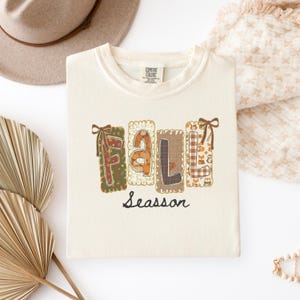 Puede incluir: Una camiseta de color crema con la palabra FALL en letras grandes y cuadradas con patrones variados que incluyen calabazas, cuadros y animales del bosque, rematadas con lazos marrones. La palabra Seasson está impresa en cursiva negra.