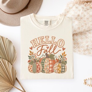 Puede incluir: Una camiseta de color crema con el texto HELLO Fall en letras marrones sobre un gráfico de tres calabazas con varios patrones, incluyendo cuadros, flores y estampados de espiga, rodeadas de hojas de otoño y pequeñas flores.