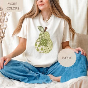 Puede incluir: Una camiseta de manga corta color marfil con un gráfico central grande de una pera compuesta por varios parches de tela verde y blanca con patrones de cuadros, flores y corazones, con un tallo marrón en la parte superior.