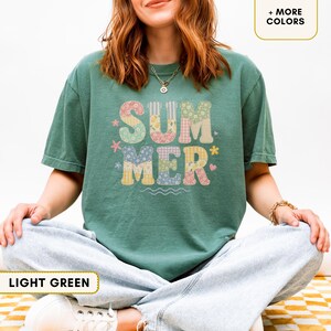 Puede incluir: Una camiseta de manga corta de color verde oscuro con la palabra SUMMER impresa en letras grandes. Cada letra está rellena con un patrón floral y de cuadros en colores pastel diferentes. Pequeños iconos de flores y corazones rodean el texto.