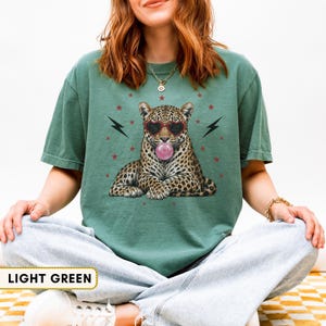 Puede incluir: Una camiseta verde claro con un gráfico de un leopardo que lleva gafas de sol en forma de corazón y hace una burbuja de chicle rosa. El diseño incluye rayos negros y pequeñas estrellas rojas que rodean al leopardo en la parte frontal.