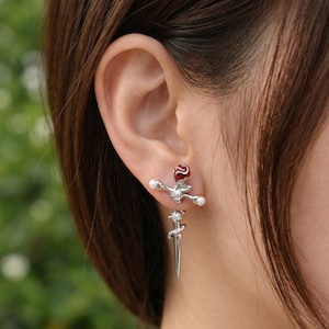 May include: A pair of silver earrings featuring a red rose and a sword design. The earrings have a post-back closure and are adorned with pearls and a small gemstone. The earrings are worn on the ear.