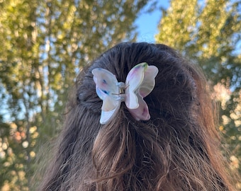 May include: A butterfly-shaped hair clip in shades of white, pink, and blue is clipped into brown hair. The clip has a gold-colored clasp and is set against a blurred background of green foliage and sunlight.