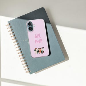 Puede incluir: Una funda de smartphone de color rosa claro con el texto GRL PWR en una fuente manuscrita sobre una peque&ntilde;a ilustraci&oacute;n gr&aacute;fica de un grupo diverso de seis personas de pie una al lado de la otra, colocada sobre un cuaderno de espiral gris oscuro.