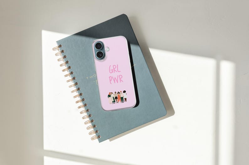 Puede incluir: Una funda de smartphone de color rosa claro con el texto GRL PWR en una fuente manuscrita sobre una peque&ntilde;a ilustraci&oacute;n gr&aacute;fica de un grupo diverso de seis personas de pie una al lado de la otra, colocada sobre un cuaderno de espiral gris oscuro.