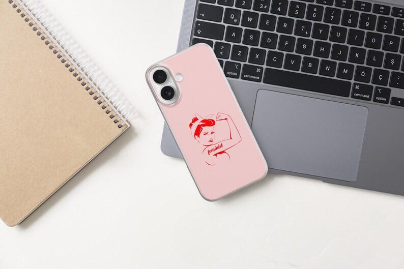 Puede incluir: Una funda de smartphone de color rosa claro con una ilustraci&oacute;n gr&aacute;fica roja de una persona flexionando el brazo con la palabra feminist escrita en cursiva. La funda tiene dos aberturas circulares para las lentes de la c&aacute;mara.