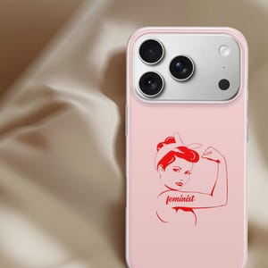 Puede incluir: Una funda de smartphone rosa claro con una ilustraci&oacute;n gr&aacute;fica roja de una persona con diadema flexionando el brazo. La palabra feminist est&aacute; impresa en letra cursiva roja en el brazo de la figura. La funda tiene un recorte para tres c&aacute;maras.