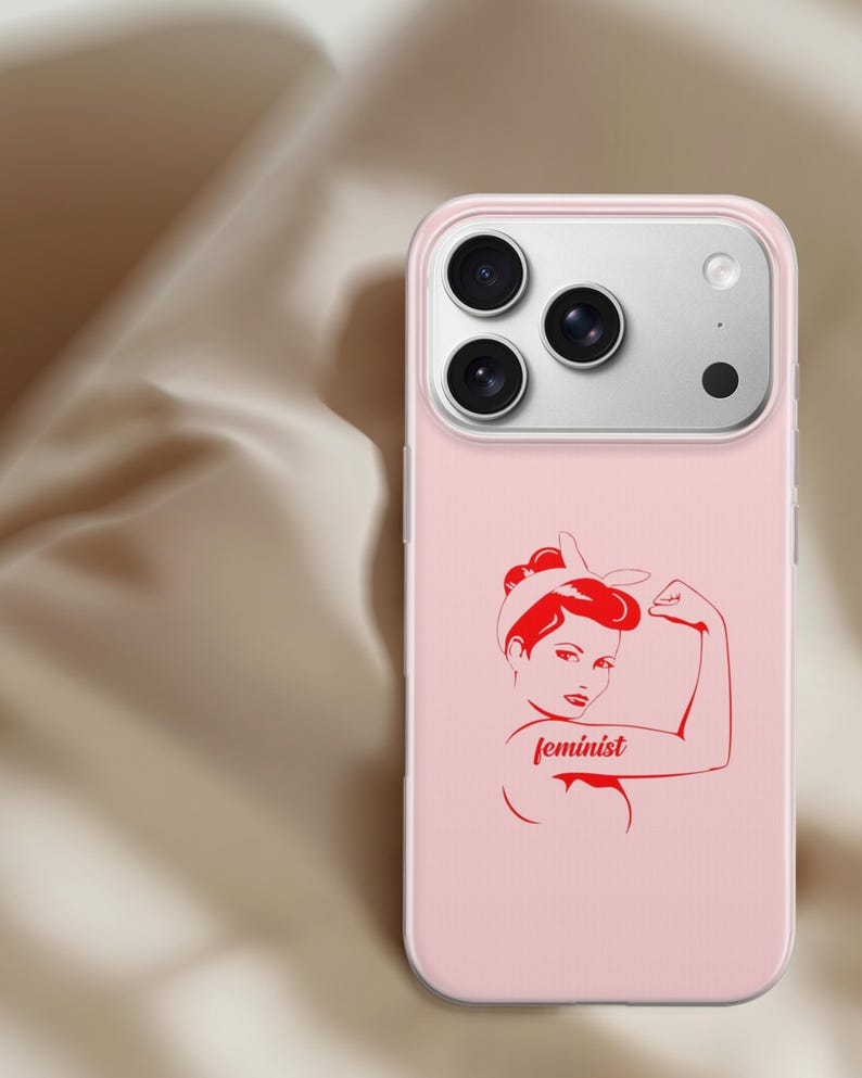 Puede incluir: Una funda de smartphone rosa claro con una ilustraci&oacute;n gr&aacute;fica roja de una persona con diadema flexionando el brazo. La palabra feminist est&aacute; impresa en letra cursiva roja en el brazo de la figura. La funda tiene un recorte para tres c&aacute;maras.